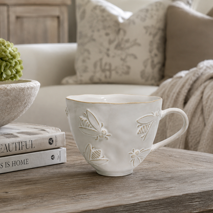 A white ceramic teacup with embossed floral and butterfly motifs sits on a rustic wooden table in a cozy living room setting, styled with books and a green hydrangea.