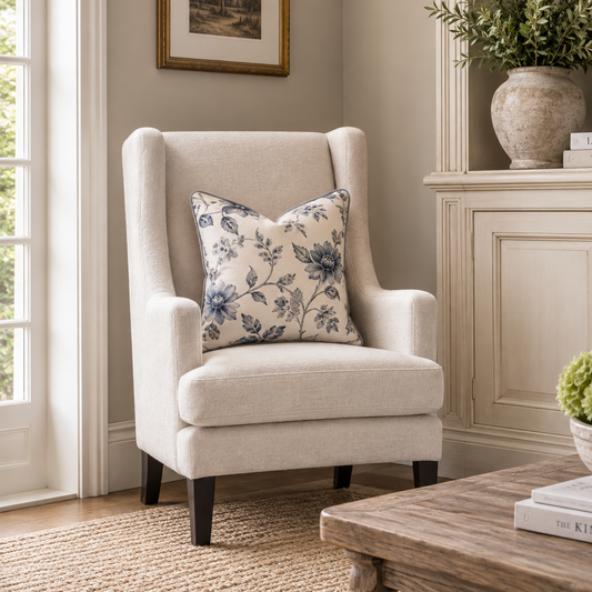 A light beige upholstered wingback armchair with dark legs, styled in a bright living room with a blue floral throw pillow, a wooden coffee table, and a cream cabinet in the background.