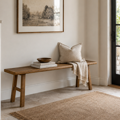 A rustic wooden entryway bench with a neutral beige throw pillow and a cream fringed throw blanket, styled with books and a small wooden bowl. Above the bench hangs a framed sepia-toned landscape print.