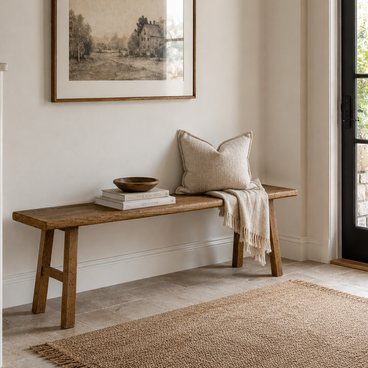 A rustic wooden entryway bench with a neutral beige throw pillow and a cream fringed throw blanket, styled with books and a small wooden bowl. Above the bench hangs a framed sepia-toned landscape print.