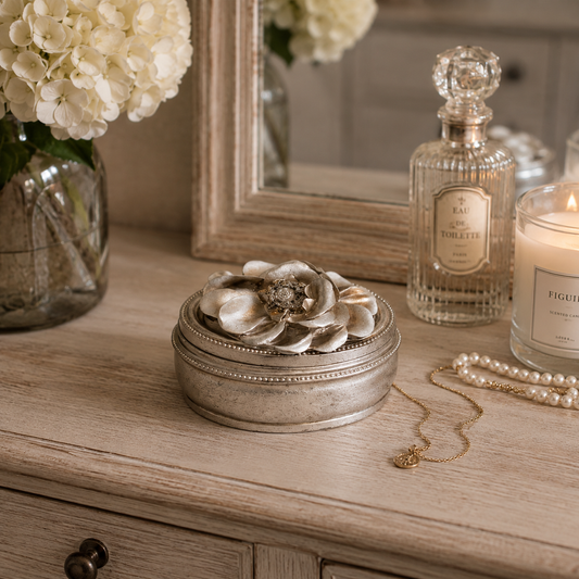 A round silver-toned jewelry box with a large flower-shaped lid ornament, styled on a wooden dresser beside a pearl necklace, a lit candle, a perfume bottle, and a vase of white flowers.