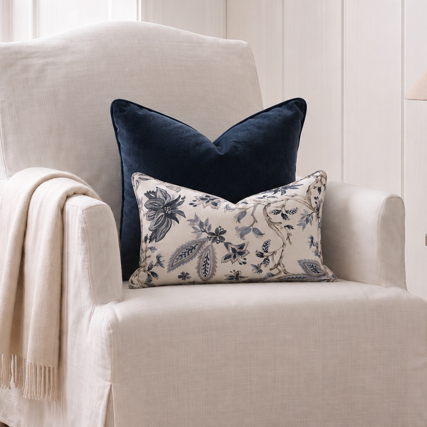 A cream slipcovered armchair in a bright room, styled with a navy blue velvet pillow, a cream floral lumbar pillow, and a draped cream throw blanket.