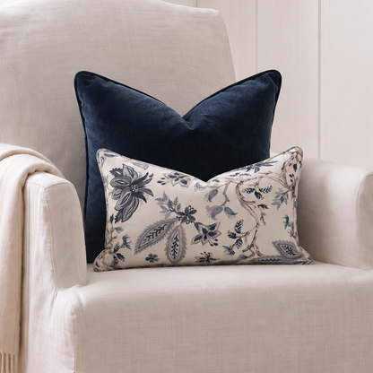 A cream slipcovered armchair in a bright room, styled with a navy blue velvet pillow, a cream floral lumbar pillow, and a draped cream throw blanket.
