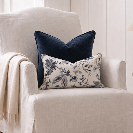 A cream slipcovered armchair in a bright room, styled with a navy blue velvet pillow, a cream floral lumbar pillow, and a draped cream throw blanket.