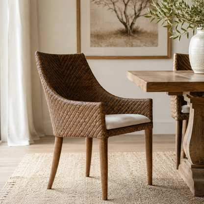 A woven wicker dining chair with a light beige cushion sits beside a rustic wooden table in a bright, modern dining room.