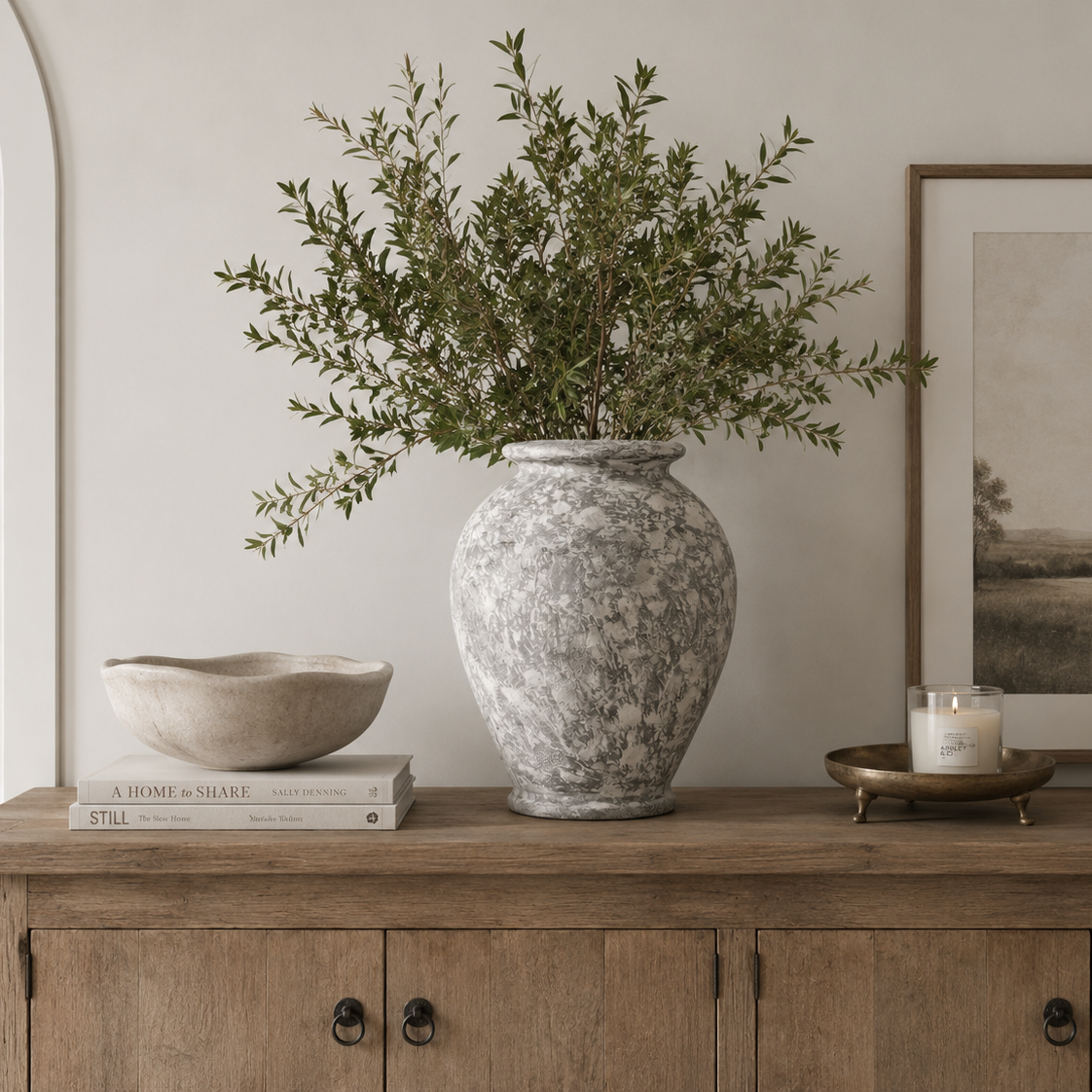 A tall textured gray and white ceramic vase filled with green foliage, styled on a wooden console table in a neutral-toned room alongside a bowl, books, a candle, and framed wall art.