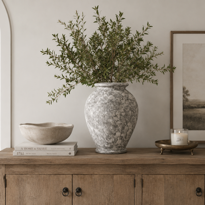 A tall textured gray and white ceramic vase filled with green foliage, styled on a wooden console table in a neutral-toned room alongside a bowl, books, a candle, and framed wall art.