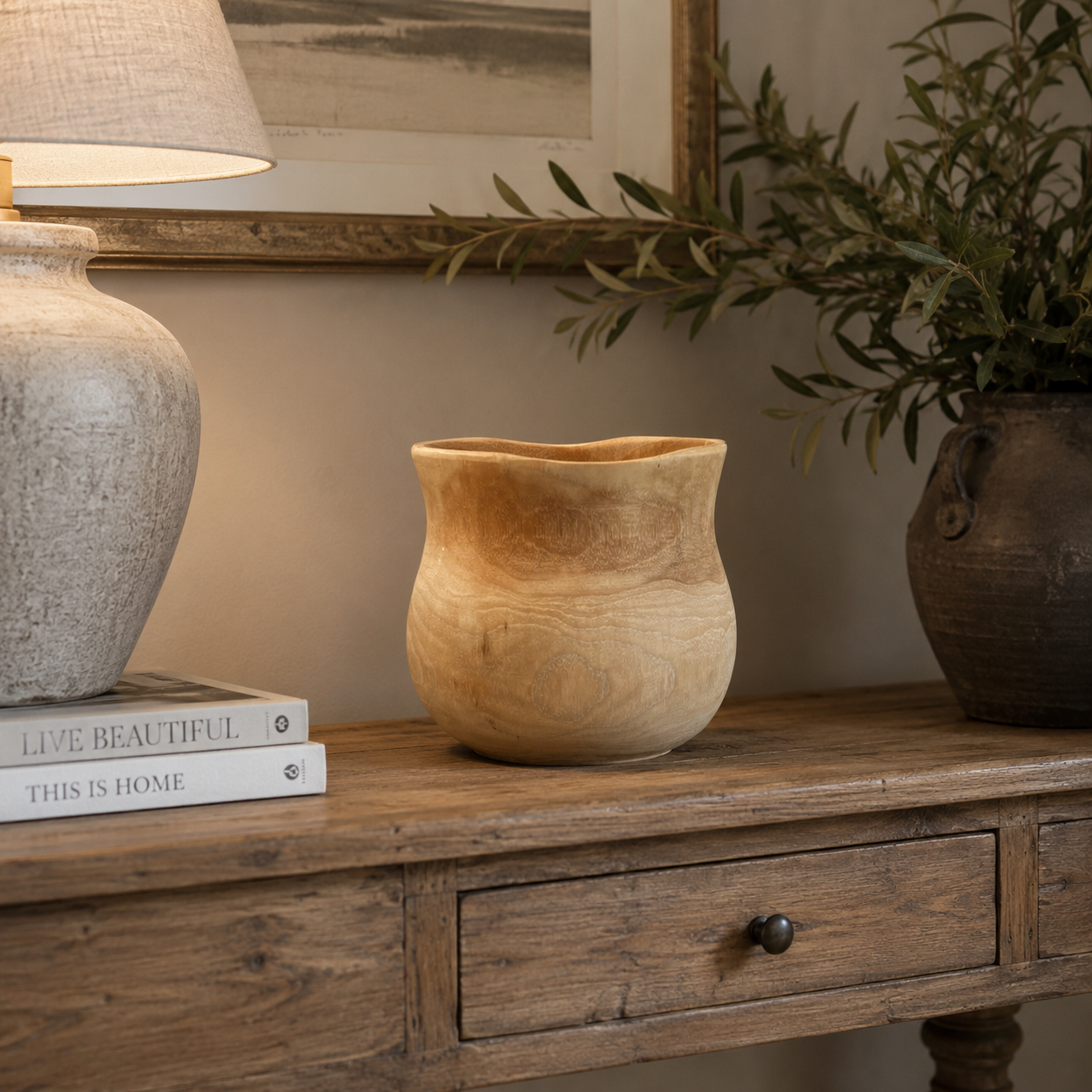 A small, rounded wooden planter with a natural light tan finish sits on a rustic wooden console table, styled with a lamp, books, and a potted olive branch in the background.