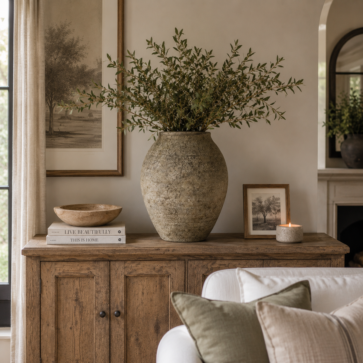 A large textured beige ceramic vase filled with green leafy branches sits on a rustic wooden sideboard in a styled living room. The scene includes framed wall art, a small wooden bowl, books, a candle, and part of a white sofa with neutral pillows.