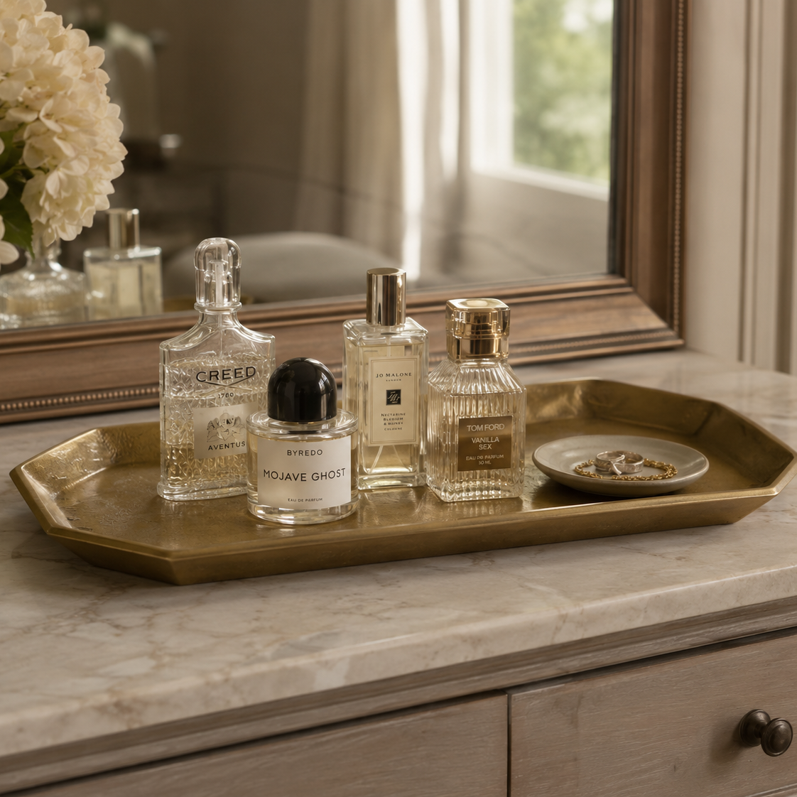 A rectangular gold-toned metal tray sits on a marble vanity top, styled with several perfume bottles and a small dish of rings. A mirror and floral arrangement are visible in the background, creating a warm, elegant interior setting.