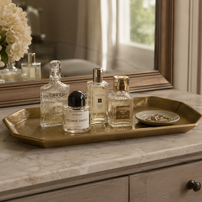A rectangular gold-toned metal tray sits on a marble vanity top, styled with several perfume bottles and a small dish of rings. A mirror and floral arrangement are visible in the background, creating a warm, elegant interior setting.