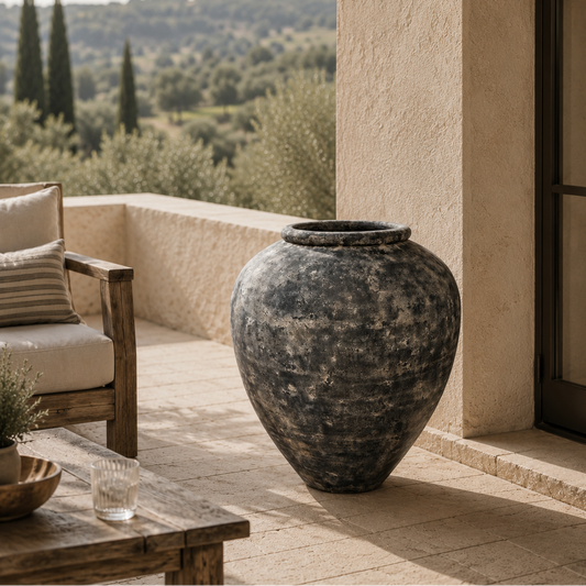A large rustic dark gray ceramic urn-style planter sits on a sunlit stone patio beside a beige stucco wall, with a wooden chair and table nearby and a scenic olive grove in the background.