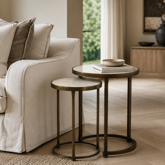 Two round nesting side tables in a warm neutral living room setting, one larger table with a marble top and one smaller stool-style table, next to a cream slipcovered sofa.