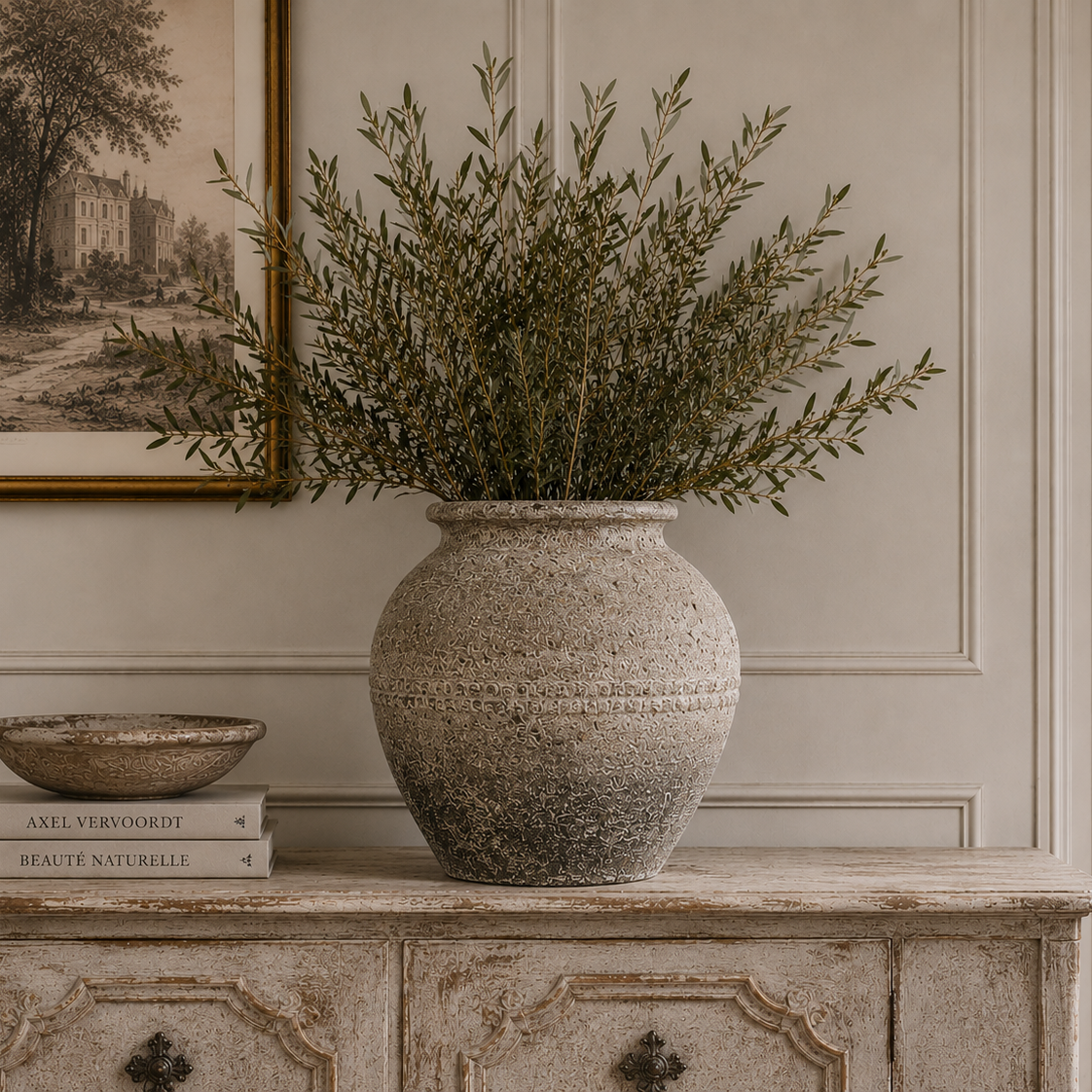 A large textured stone-style vase filled with green foliage sits on a distressed wooden console table in a styled interior room. A framed print, books, and a small bowl are also visible on the table.