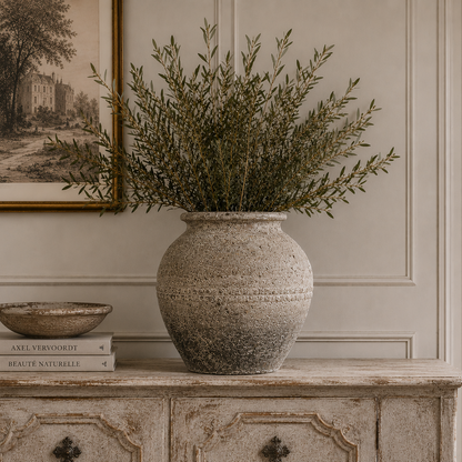 A large textured stone-style vase filled with green foliage sits on a distressed wooden console table in a styled interior room. A framed print, books, and a small bowl are also visible on the table.