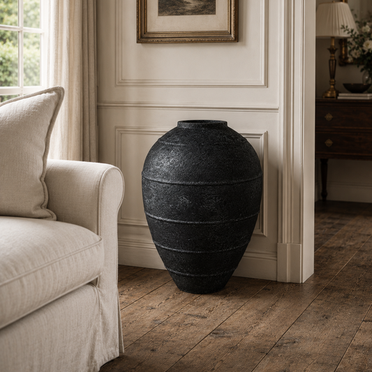 A large, textured black ceramic vase with horizontal ridges, displayed in a classic living room setting next to a cream-colored armchair on a rustic wooden floor.