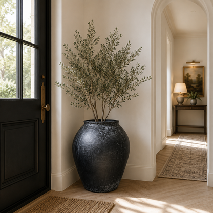 A large black textured ceramic planter holding a tall olive branch arrangement, placed in a bright entryway next to a black door, with a hallway and rug visible in the background.