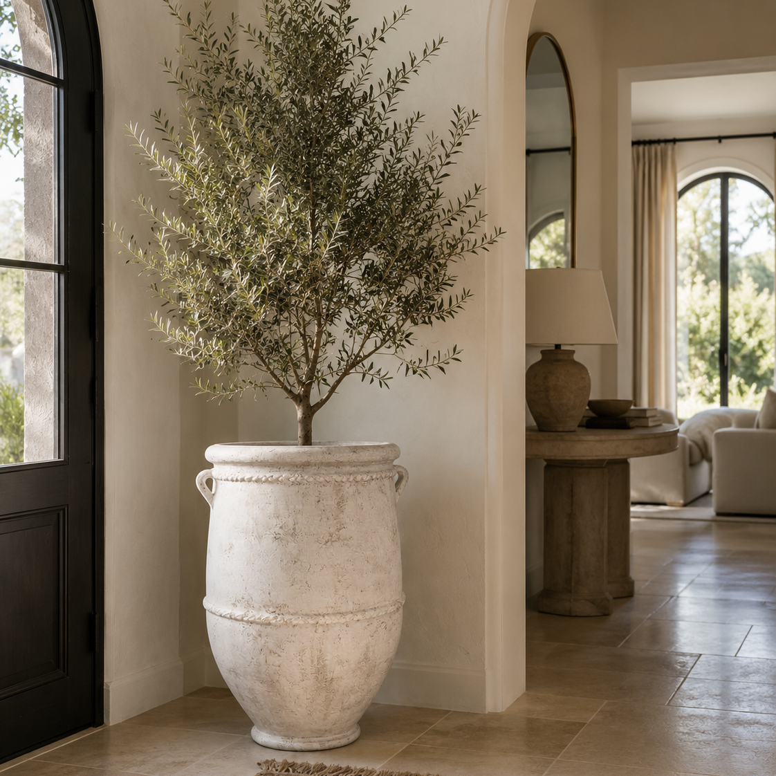 A large potted olive tree in a distressed white ceramic urn-style planter, placed in a bright, elegant entryway with beige walls, tiled floors, and an arched doorway in the background.