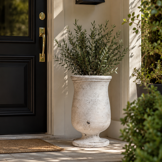 A large distressed white stone urn planter filled with green foliage, placed on a stone doorstep next to a black front door with gold hardware.