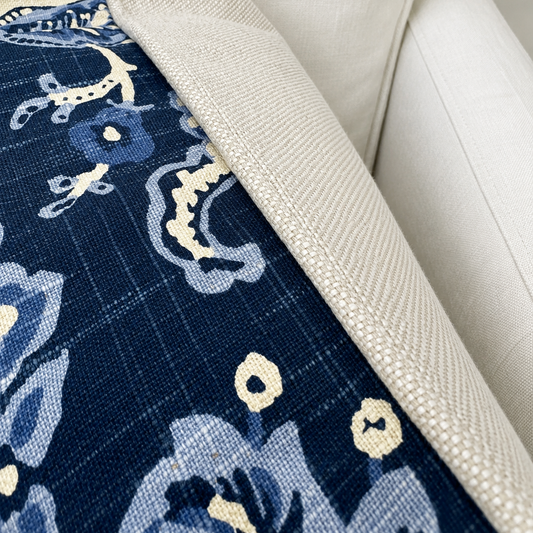 Close-up of a navy blue patterned textile, likely a throw blanket or cushion cover, draped over a light beige upholstered chair. The textile features a floral and nautical-inspired print in navy, light blue, and cream, with a textured cream border.