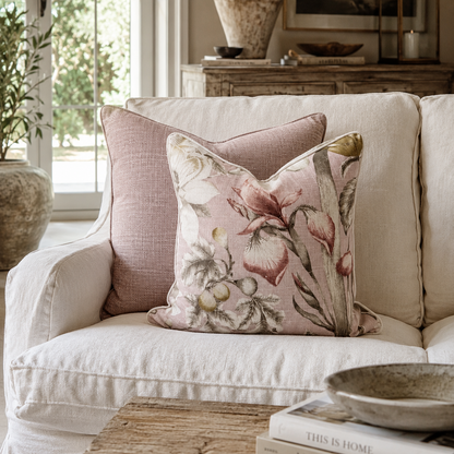 A styled interior scene showing a cream upholstered sofa with two decorative throw pillows. The front pillow is square and features a soft pink floral print with green and beige botanical details.