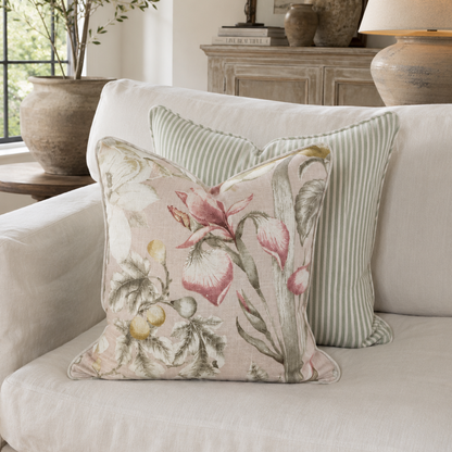 A square decorative throw pillow with a pink and green floral pattern sits on a white sofa in front of a striped pillow, in a styled living room setting.