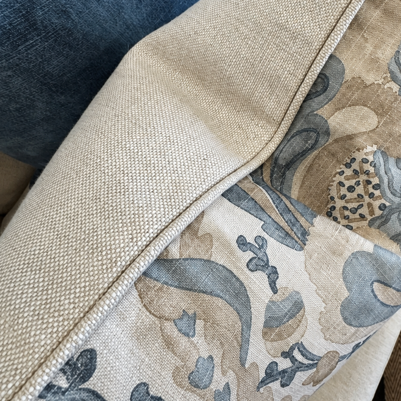 A close-up of a decorative throw pillow with a beige and blue-gray abstract pattern, resting on a light beige sofa next to a blue cushion.