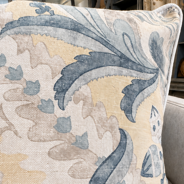 A square throw pillow with a blue and beige botanical pattern, resting on a light gray fabric sofa in a living room setting.