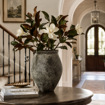 A rustic gray stone vase filled with faux magnolia flowers and green leaves, displayed on a round wooden table in a bright, elegant interior hallway.