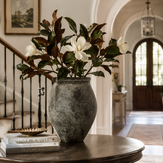 A rustic gray stone vase filled with faux magnolia flowers and green leaves, displayed on a round wooden table in a bright, elegant interior hallway.