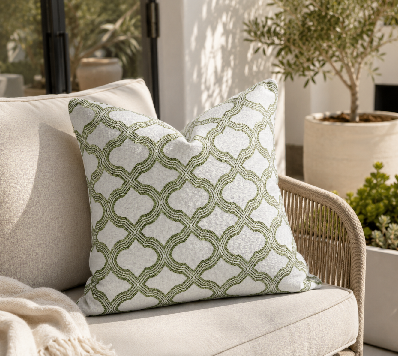 A square throw pillow with an olive green trellis pattern on a white background, styled on a beige outdoor sofa next to a wicker chair in a sunlit patio setting.