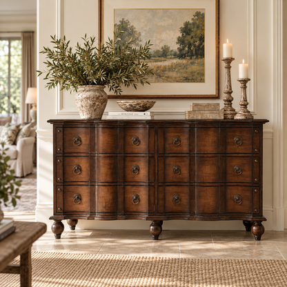 A dark brown wooden chest of drawers with multiple drawers and round metal knobs, styled in a classic living room setting with a potted olive branch, books, candles, and a framed landscape painting above it.