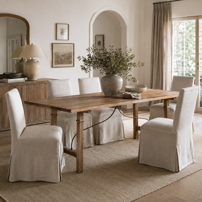 A rustic wooden dining table with four upholstered slipcovered chairs in a bright, neutral-toned dining room. The scene includes a large ceramic vase with greenery, a sideboard with lamps, framed wall art, and a window with curtains.