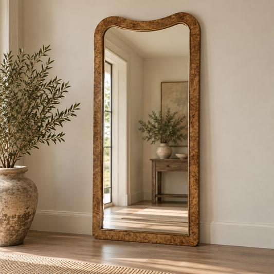 A tall floor mirror with a textured, wavy gold-brown frame stands on a light wood floor against a beige wall, reflecting a softly lit interior room with a console table and potted plants.
