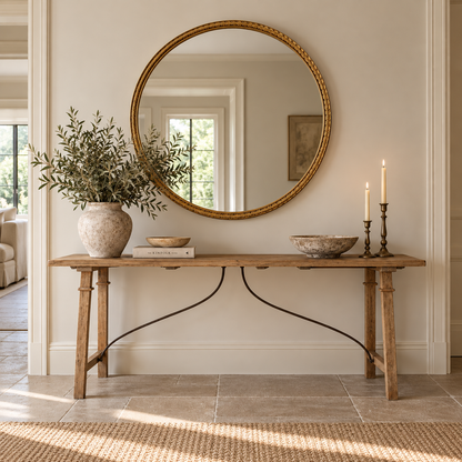 A round gold-framed mirror hangs on a beige wall above a rustic wooden console table styled with a large ceramic vase of greenery, books, bowls, and two lit candlesticks in a bright, sunlit room.