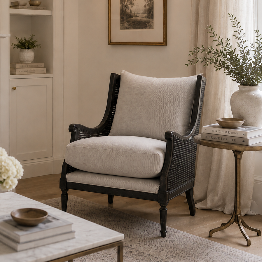 A styled interior scene featuring a black wicker armchair with light beige cushions, positioned next to a marble coffee table, a gold side table, and a white fireplace mantel in a bright, elegant room.