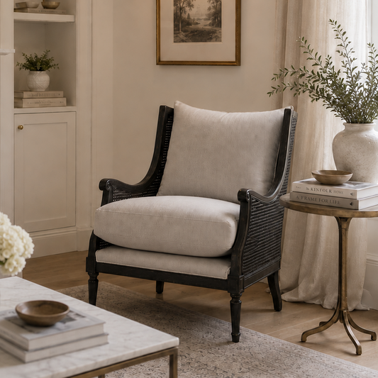 A styled interior scene featuring a black wicker armchair with light beige cushions, positioned next to a marble coffee table, a gold side table, and a white fireplace mantel in a bright, elegant room.