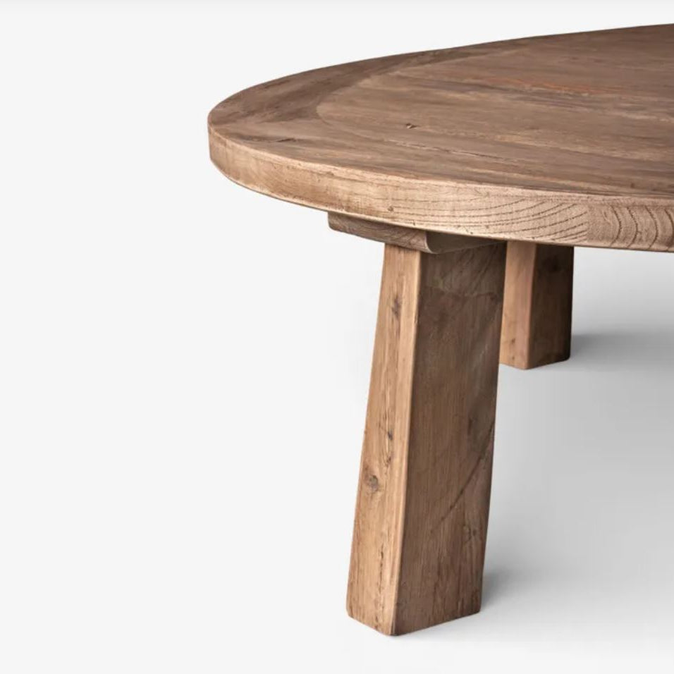 A close-up of a round wooden table with a thick, rustic leg base, shown against a plain light gray background.