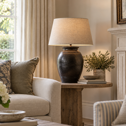 A rustic dark brown textured ceramic table lamp with a beige linen drum shade sits on a wooden side table in a beautifully styled living room next to a cream sofa.