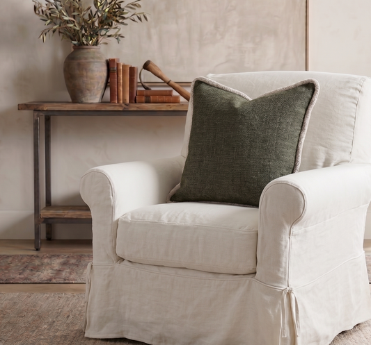 White armchair with a green pillow in a room with a wooden side table and plant.