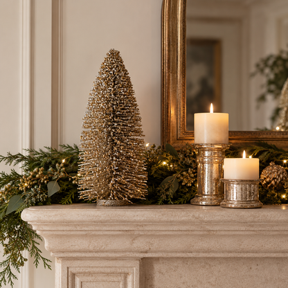A decorative gold wire Christmas tree stands on a stone mantel beside lit white pillar candles in silver holders, with green garland and a mirror in the background.
