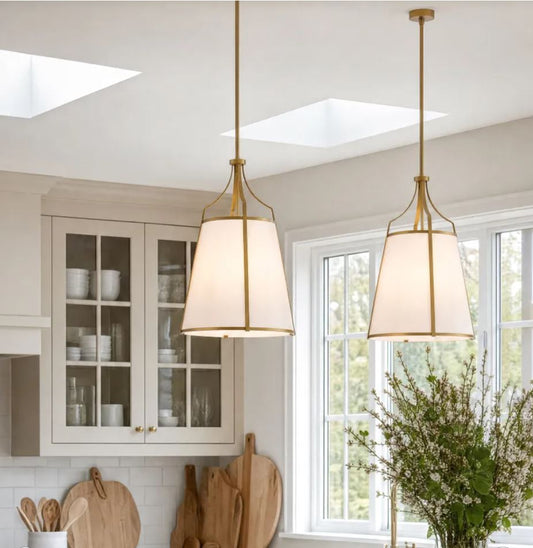 Two brass pendant light fixtures with white drum shades hanging in a bright, modern kitchen with white cabinets, windows, and a potted plant.