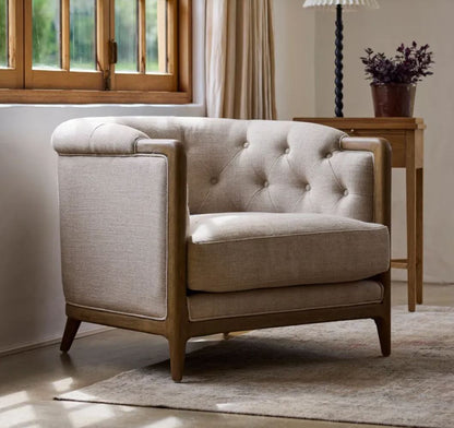 A light beige upholstered armchair with a tufted backrest and wooden legs, shown in a styled living room setting with a side table, lamp, window, and rug.