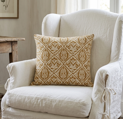 White armchair with a patterned pillow in a room with a wooden table and framed picture.
