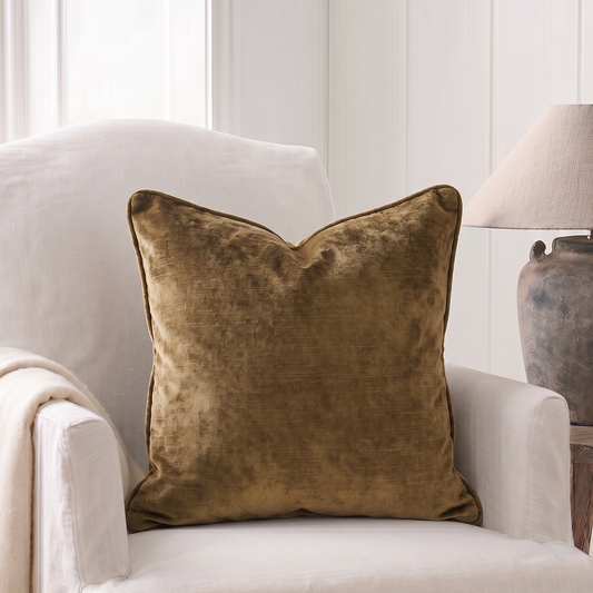 A square throw pillow in a textured burnished gold  velvet fabric, styled on a white armchair next to a wooden side table with a lamp.