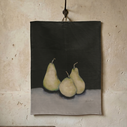Tea towel with a painting of three pears on a dark background, hanging against a textured wall.