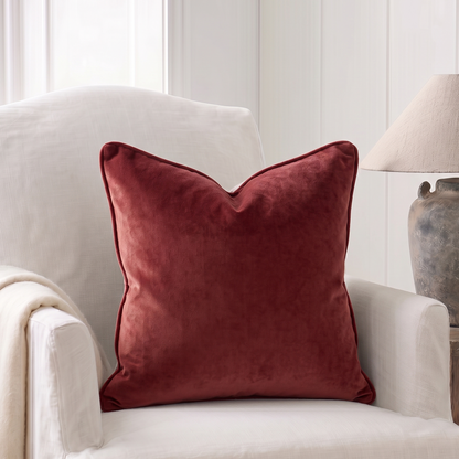 A square deep vintage rose 
velvet throw pillow with piped edges sits on a cream upholstered armchair in a bright, neutral-toned living room setting.