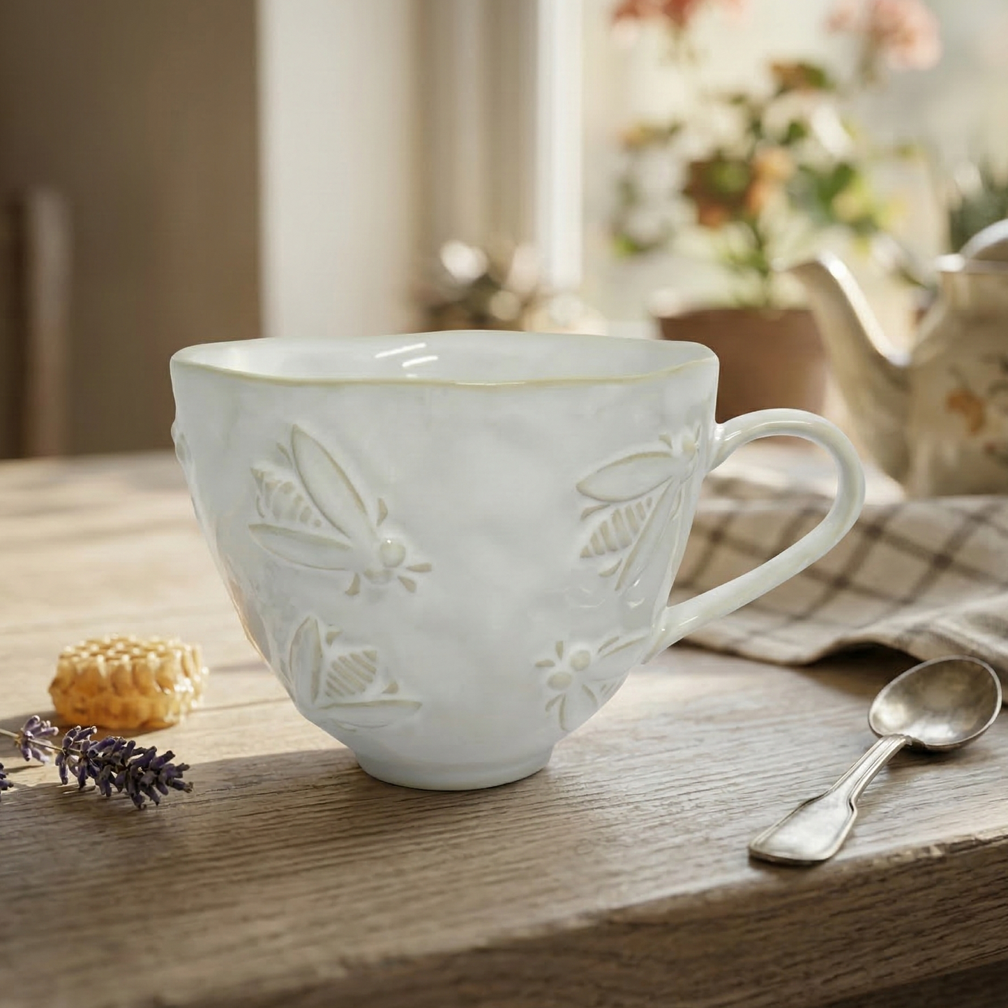White ceramic mug with bee embossing on a wooden table with a teapot and lavender.