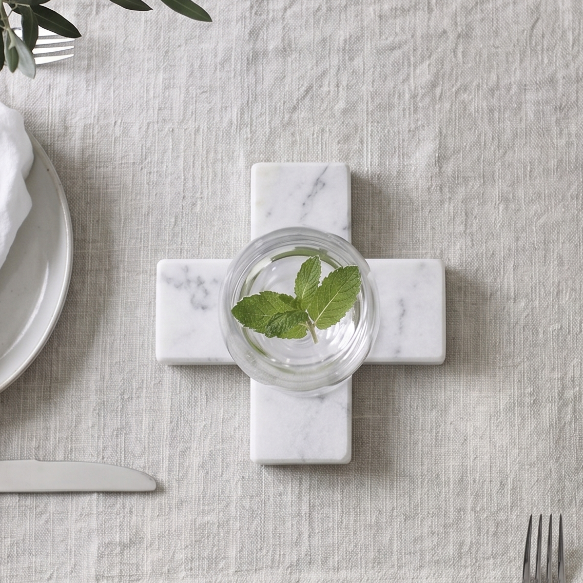 A styled tabletop setting featuring a light beige linen table runner laid across a gray wood-grain table. In the center of the runner sits a white marble cross-shaped coaster holding a small glass of water with green mint leaves.