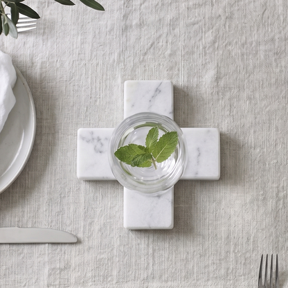 A styled tabletop setting featuring a light beige linen table runner laid across a gray wood-grain table. In the center of the runner sits a white marble cross-shaped coaster holding a small glass of water with green mint leaves.